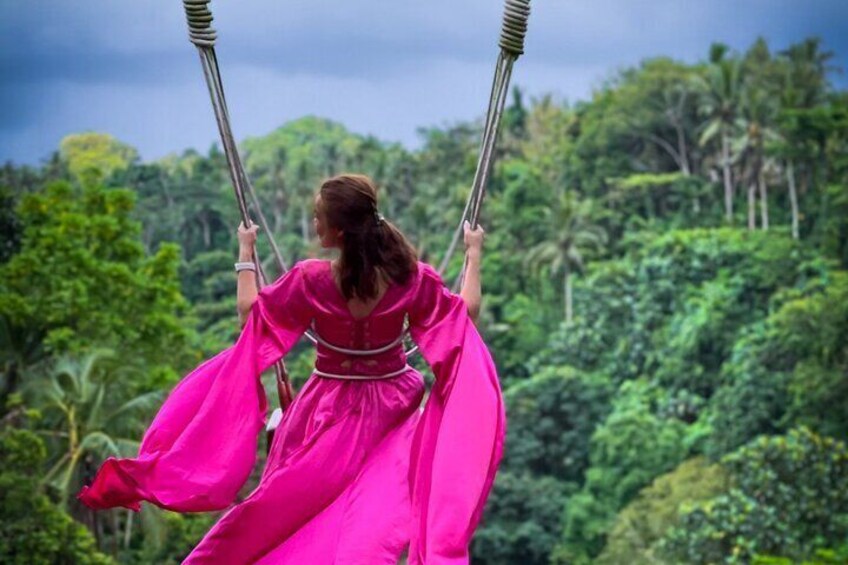 A person swinging on a swing with a pink dress, surrounded by lush green foliage. 3