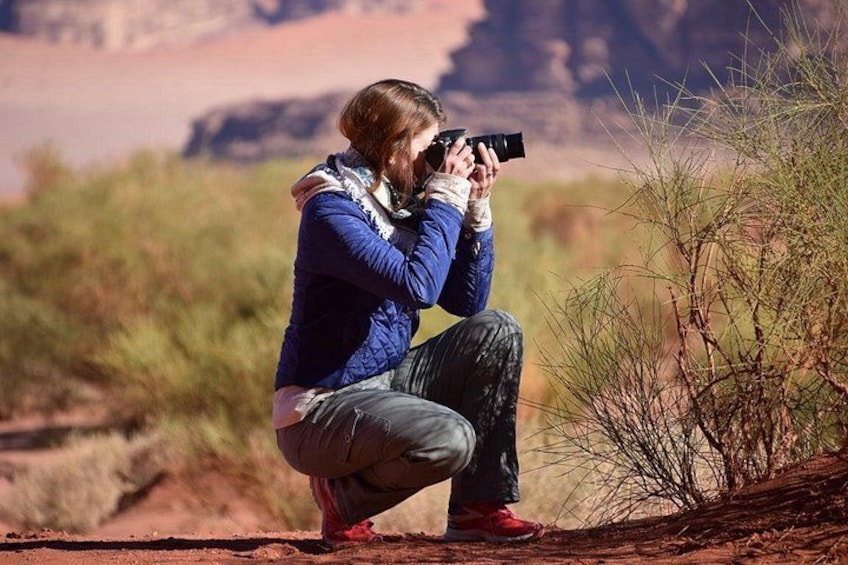 A person crouching and taking a photograph in a desert landscape with sparse vegetation and rocky terrain. 3