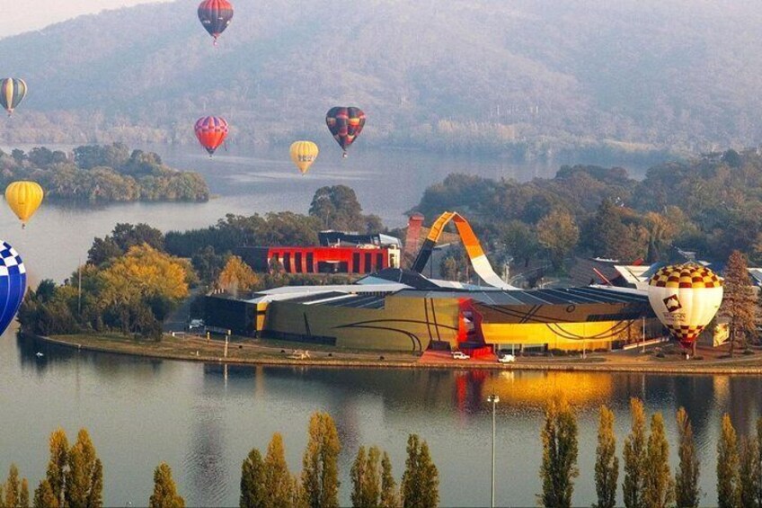 A hot air balloon festival with multiple balloons over a lake and a large hangar. 2