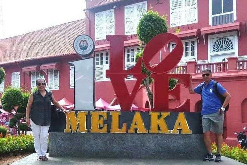 Ein großes Schild mit der Aufschrift 'LOVE MELAKA' in Rot und Gelb, im Hintergrund ein Gebäude mit roten Wänden und weißen Fenstern sowie ein kleines, kreisförmiges Emblem auf dem Schild. 1