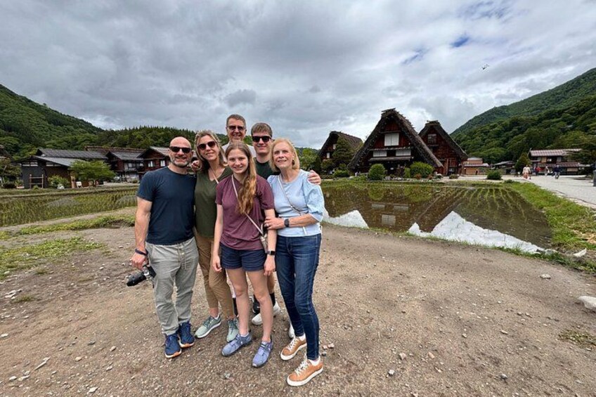 En gruppe mennesker står foran traditionelle huse med en sø og bjerge i baggrunden. 4