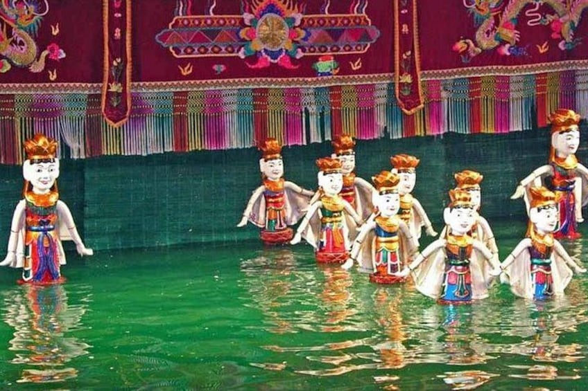 Eine Gruppe von Personen in traditioneller Kleidung steht im Wasser vor einem farbenfrohen Hintergrund. 2