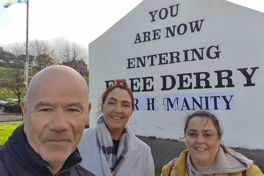 Three people in front of a sign that reads 'YOU ARE NOW ENTERING FREE DERRY FOR HUMANITY.' 3