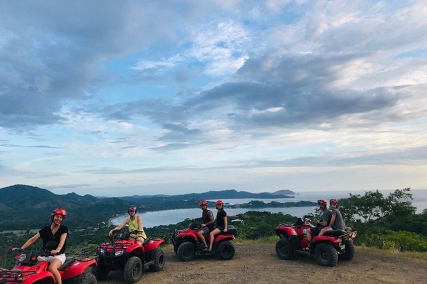 Three people on red ATVs on a dirt path with a scenic view of mountains and a body of water in the background. 1