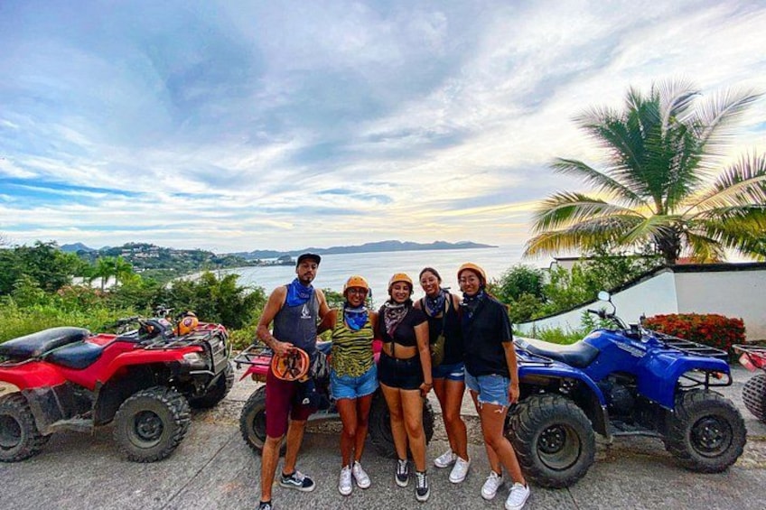 A group of people posing with ATVs in a tropical setting with a view of the ocean and palm trees. 4