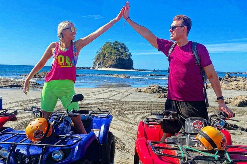 Two people high-fiving on a beach with ATVs and a clear blue sky. 3