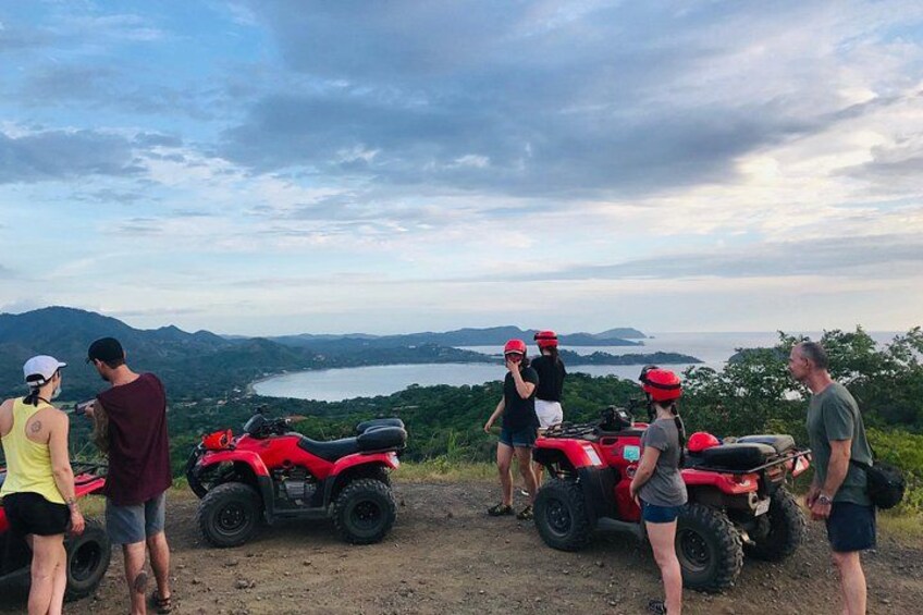 A group of people on a dirt path with red all-terrain vehicles, overlooking a scenic landscape with hills and a body of water. 5