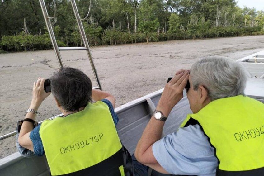 Two individuals wearing high-visibility vests on a boat, with a sandy beach and trees in the background. 4