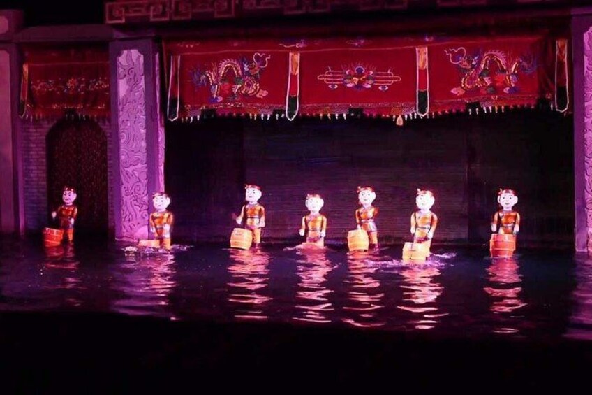 A stage with performers in traditional costumes, a red backdrop with dragon motifs, and a water feature. 2