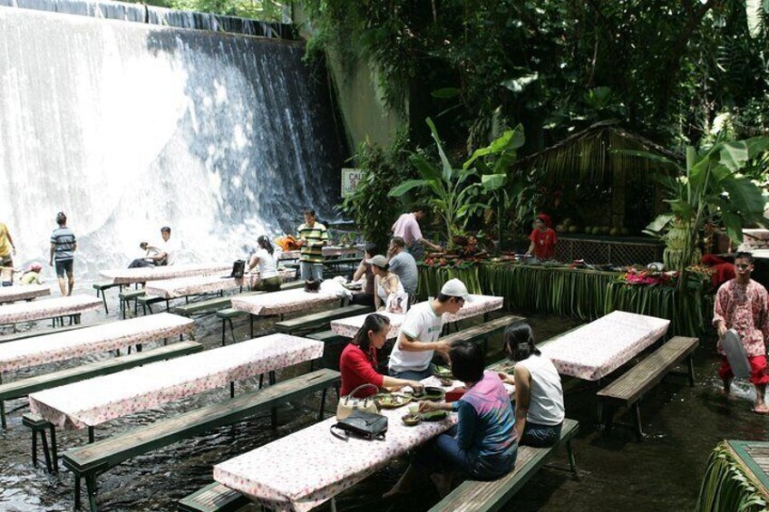 People dining at tables near a waterfall in a lush, green setting. 1