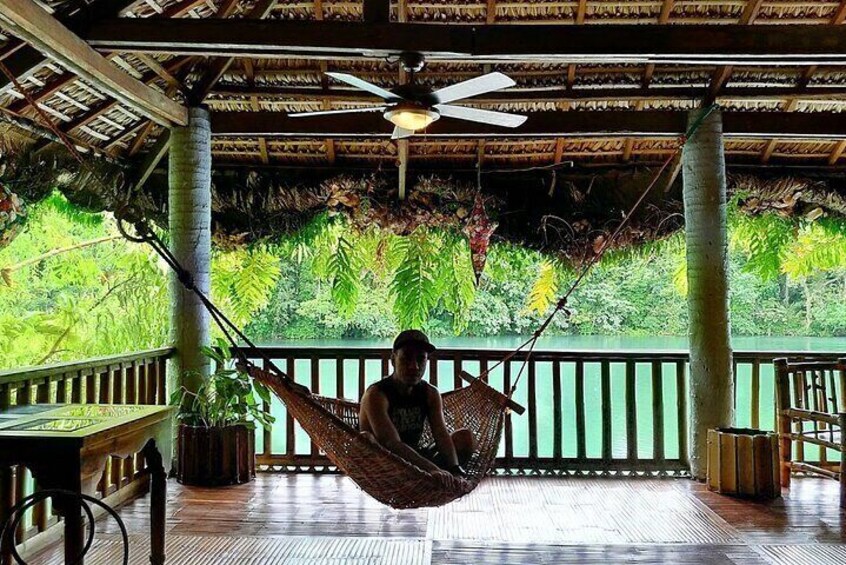 A person relaxing in a hammock on a wooden deck with a thatched roof, surrounded by lush tropical vegetation. 2
