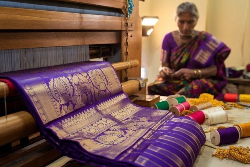 A purple fabric with a paisley pattern on a wooden loom, with spools of thread and a person working in the background. 1