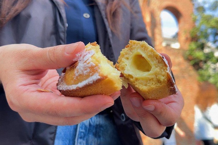 Une personne tenant à la main une pâtisserie à moitié mangée, dont la garniture est visible, portant une veste sombre, et avec une structure arquée en arrière-plan. 1