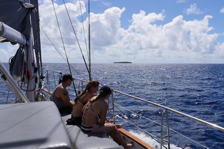 A sailboat with three people on deck, a clear sky, and a distant island. 3