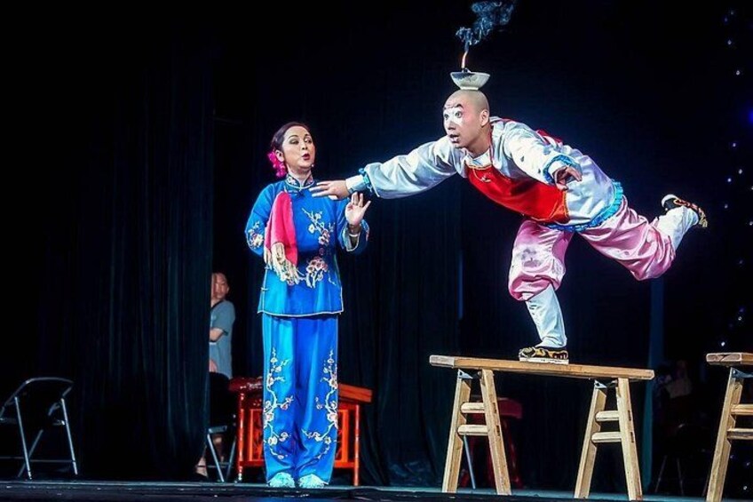A performer balancing on a stool with a hat on his head, another person in traditional blue attire, and a stage with a dark backdrop. 2