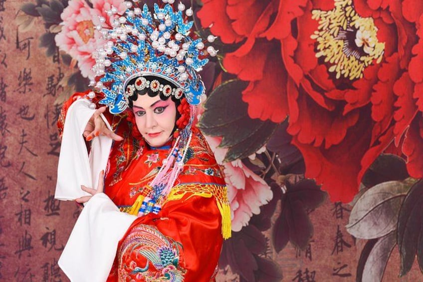 A person in traditional red and blue attire with elaborate headgear, standing in front of a floral backdrop with Chinese calligraphy. 2