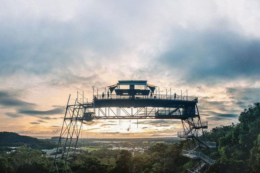 A large metal structure with a viewing platform, surrounded by trees and hills, under a cloudy sky. 2
