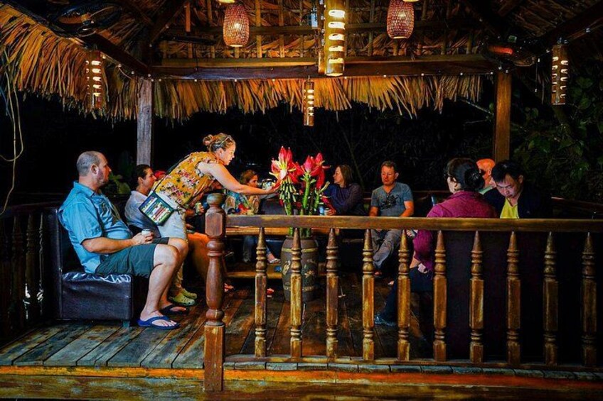 A wooden deck with people seated, a woman serving drinks, and a thatched roof structure. 4
