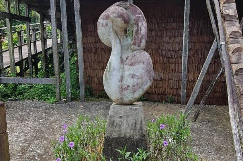 A large abstract sculpture in a garden with a wooden fence and a thatched-roof structure in the background. 2