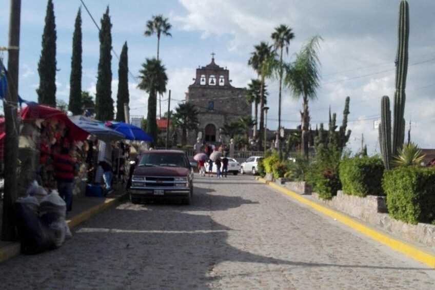 A cobblestone street with a vintage car, palm trees, cacti, and a historic building in the background. 2