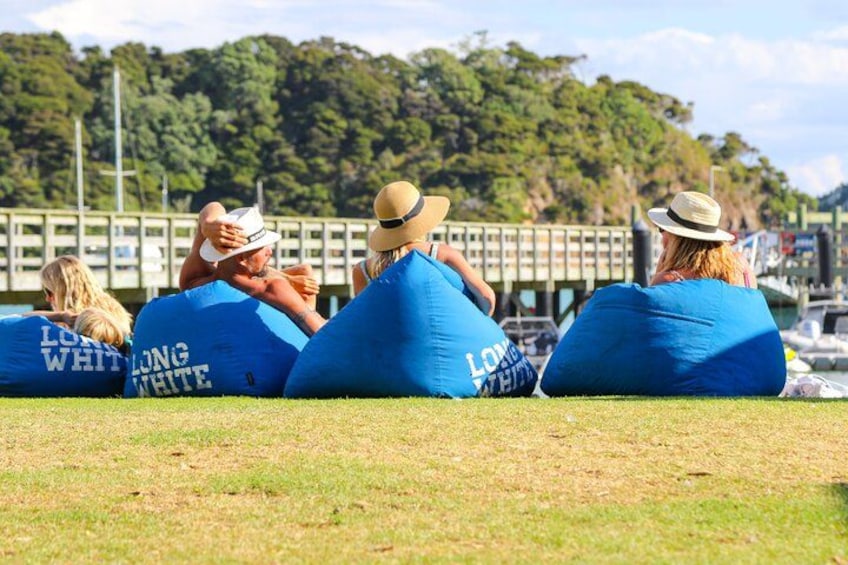 Cuatro personas sentadas en pufs azules con el texto 'LONG WHITE', al aire libre con un puente y follaje de fondo. 4