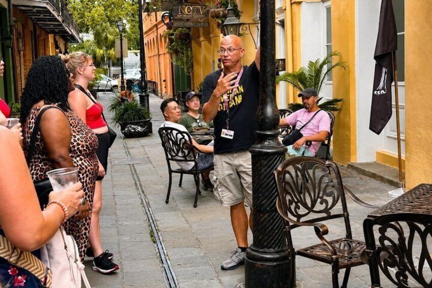 A street scene with people sitting at outdoor tables, a man speaking to a group, and a building with a sign. 5