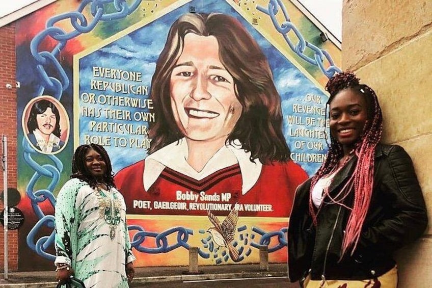 A mural with a portrait and text, two people standing in front of it, and a chain-link fence in the background. 3