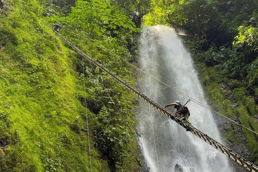 A person crossing a rope bridge over a waterfall in a lush green forest. 2