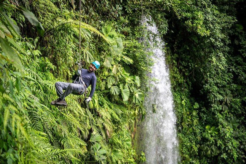 En person åker på en zipline genom en frodig skog med ett vattenfall i bakgrunden. 5