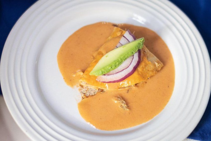 A white plate with a piece of food covered in a creamy sauce, topped with a slice of avocado and a slice of red onion. 5
