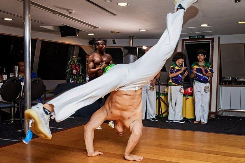 A person performing a handstand with a leg extended upwards, surrounded by other individuals in a room with a wooden floor and exercise equipment. 3