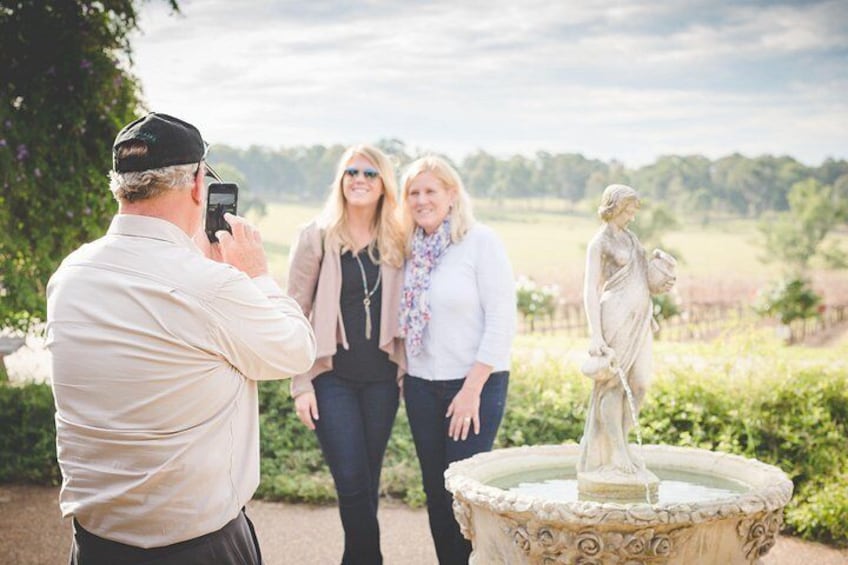 A person taking a photo of two people standing near a statue in a garden with a fountain. 3