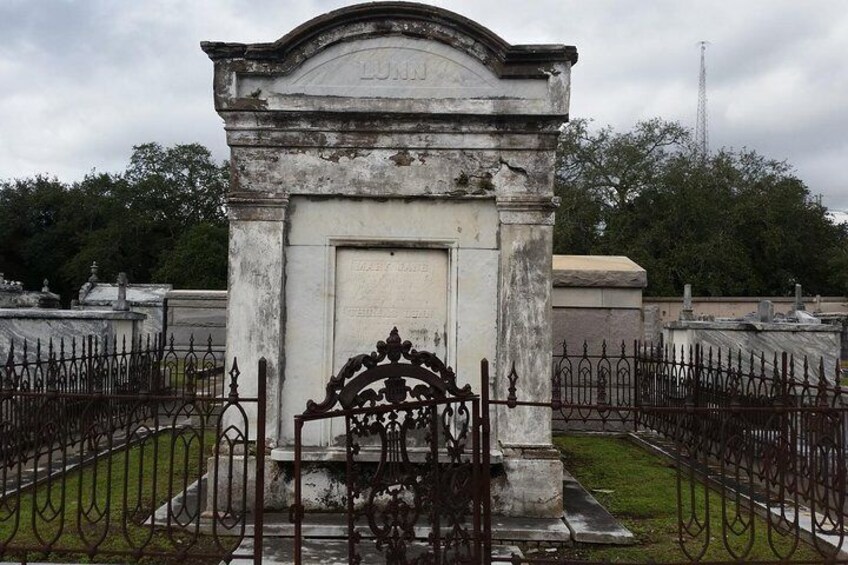 Una lápida desgastada con inscripciones, rodeada por una cerca de hierro forjado en un cementerio. 5