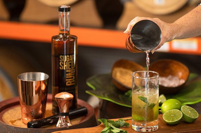 A person pouring a drink from a copper shaker into a glass, with a bottle labeled 'SUGAR SHED' and lime wedges on the table. 3