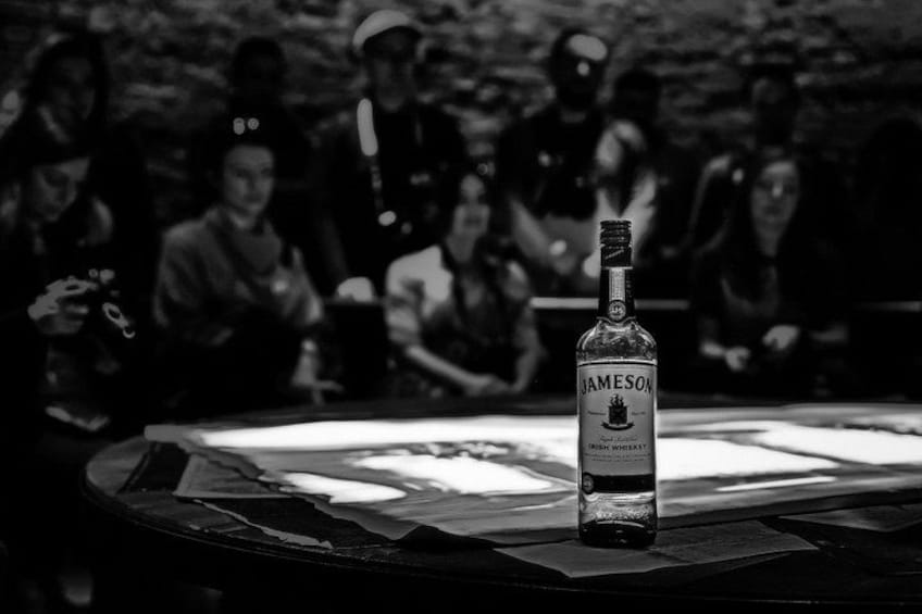 Une bouteille de whisky irlandais Jameson est posée sur une table, et on aperçoit un groupe de personnes à l’arrière-plan. 2