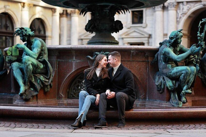 A couple sitting on a bench in front of a fountain with statues. 4