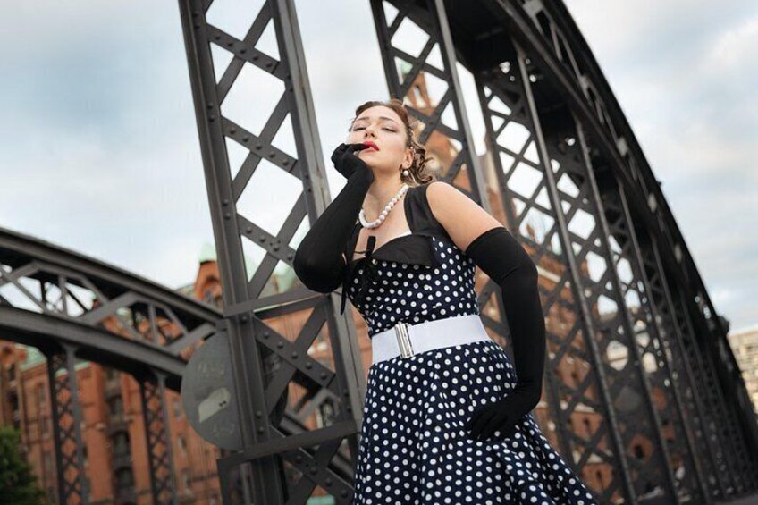 Eine Person in einem Punktekleid steht auf einer Brücke mit Metallträgern und einem Backsteingebäude im Hintergrund. 4