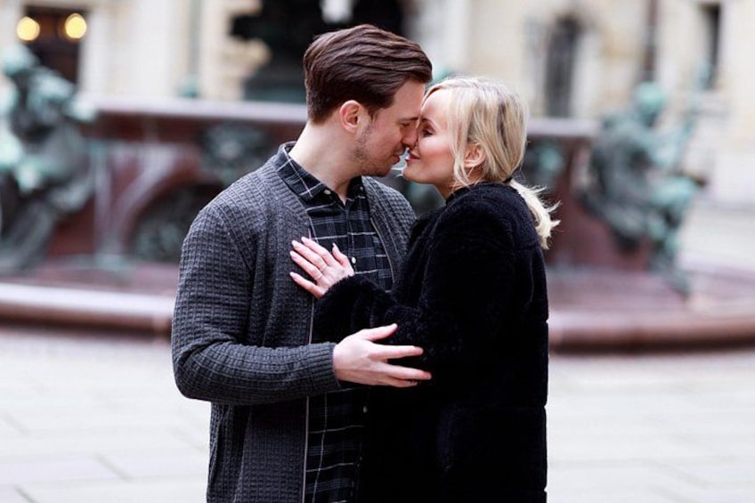 A couple embracing in an urban setting with buildings and a fountain in the background. 1