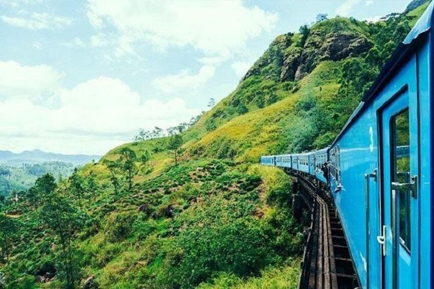 A blue train on a railway track winding through a green, hilly landscape. 1
