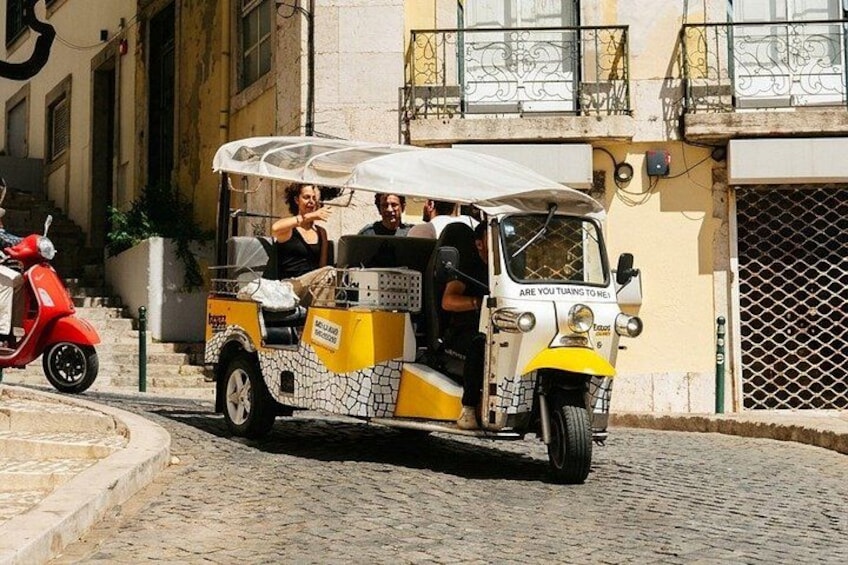 A yellow and white tuk-tuk with passengers, parked on a cobblestone street. 4