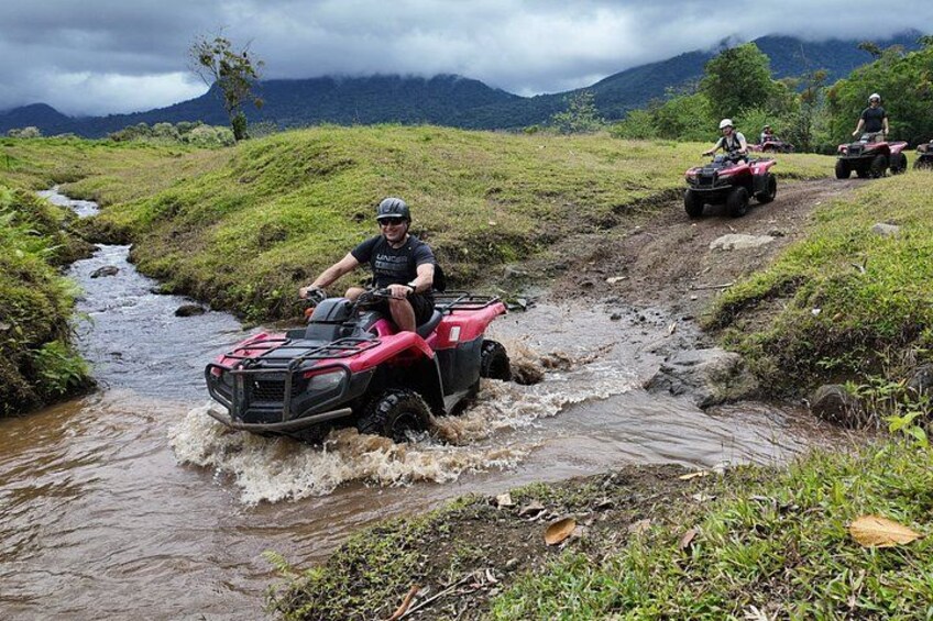 Una persona guida un quad rosso attraverso un ruscello fangoso in una zona erbosa, con montagne sullo sfondo. 2