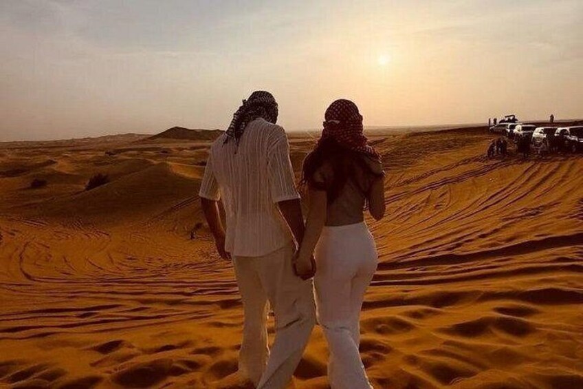 A couple walking on a desert dune, a sunset in the background, and a vehicle in the distance. 4
