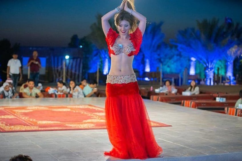 A belly dancer performing on a stage with an audience seated in the background. 1