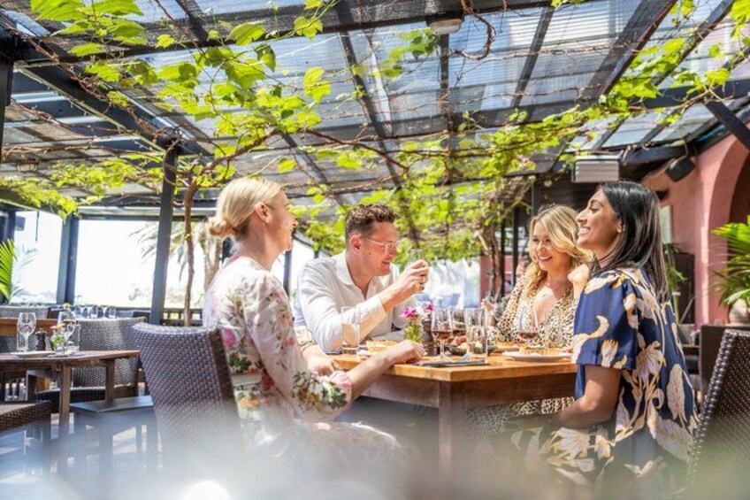 En gruppe mennesker spiser på en udendørs restaurant med glasloft og grønne planter. 1