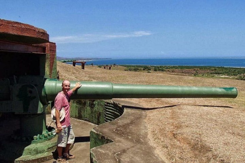 A man standing next to a large cannon, with a coastal landscape in the background. 4