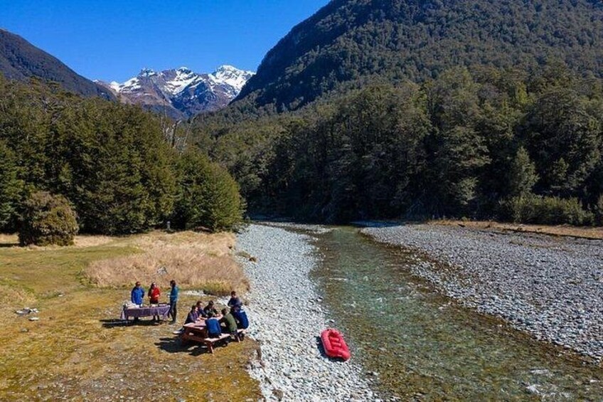 Ein Fluss mit Menschen, die auf einer Decke sitzen, einem roten Rucksack und einer bergigen Landschaft im Hintergrund. 2