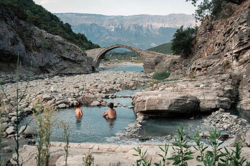 Un lecho rocoso de río con gente bañándose, un puente de piedra y montañas al fondo. 2