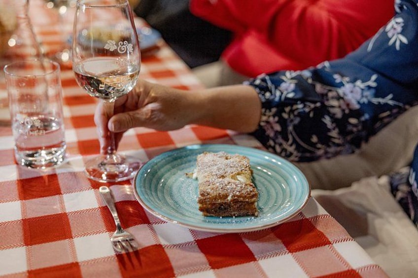 A person holding a glass of white wine, a blue plate with a dessert, and a fork on a checkered tablecloth. 5
