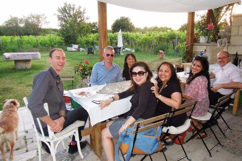 Un grupo de personas sentadas en una mesa al aire libre con mantel blanco, rodeadas de follaje y un banco de piedra. 3