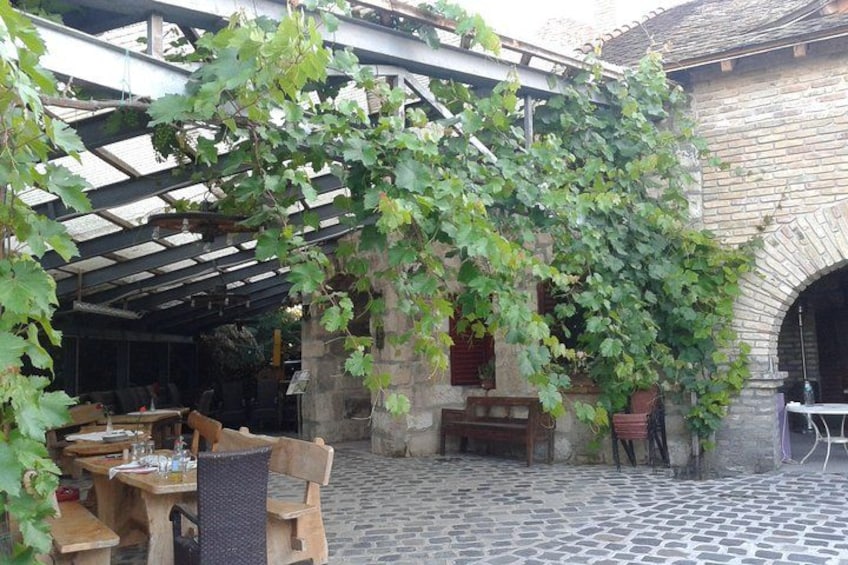 Un patio empedrado con mesas y sillas de madera, un edificio de ladrillo y una pérgola cubierta de hiedra verde. 4